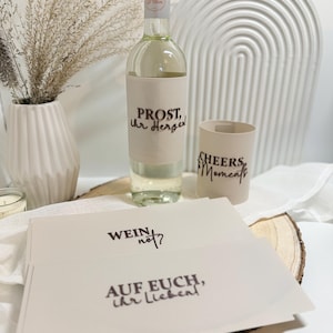 May include: A bottle of white wine with a cream-coloured label that reads "PROST, ihr Herzen!" is displayed. A matching cup with "CHEERS to Moments" is next to the bottle. Several cards with text are in front of the bottle. The theme is wine and celebration.