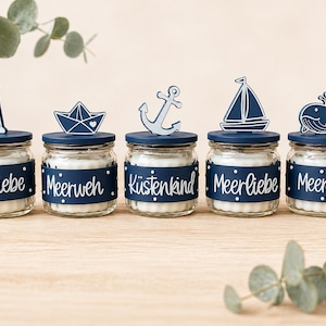 Könnte beinhalten: Fünf Gläser mit marineblauen Deckeln und Etiketten, die jeweils mit einem maritimen Holzausschnitt versehen sind. Die Etiketten lauten "Strandliebe", "Meerweh", "Küstenkind", "Meerliebe" und "Meergeküst". Die Ausschnitte umfassen einen Leuchtturm, ein Papierboot, einen Anker, ein Segelboot und einen Wal.