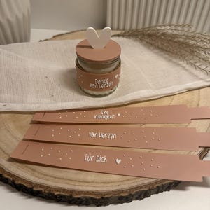 Puede incluir: Una pequeña vela en tarro de cristal con tapa en forma de corazón y una banda decorativa que dice "Danke von Herzen". También hay tres etiquetas de regalo rosas a juego con texto blanco y detalles de corazón.