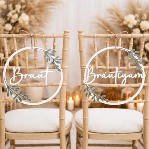 Può includere: Due sedie in legno con cuscini bianchi sono adornate da cartelli circolari. I cartelli recitano "Braut" e "Bräutigam" in elegante scrittura, decorati con accenti di foglie verdi. I cartelli sono sospesi da spago.