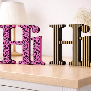 Könnte beinhalten: Zwei Holzdekorationen in Form des Buchstabens "H". Eine ist rosa mit einem schwarzen Leopardenmuster, die andere ist schwarz mit goldenen vertikalen Streifen. Sie werden auf einer hellen Holzoberfläche ausgestellt.