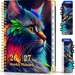 May include: A spiral-bound weekly planner featuring a vibrant, colorful illustration of a cat's face. The cat's fur is depicted in shades of blue, purple, orange, and green. The planner includes the dates "26 Jul 27 Jun" and the words "Weekly Planner."