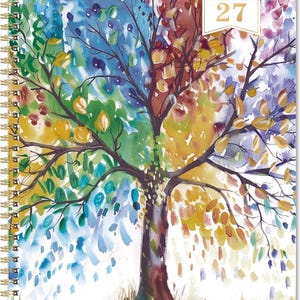May include: A spiral-bound planner with a watercolor painting of a tree representing the four seasons. The tree has a brown trunk and branches with leaves in shades of green, yellow, orange, and blue. The text "26 MONTHLY PLANNER 27" is in a gold rectangle.