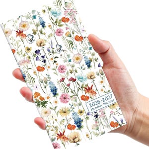 May include: A floral-patterned 2026-2027 monthly planner held in a hand. The planner features a white background with colorful flowers in shades of orange, pink, blue, and yellow. The text "2026-2027 Monthly Planner" is visible.