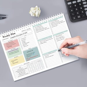 May include: A spiral-bound weekly planner with a white background, featuring sections for goals, to-do lists, and a habit tracker. The planner includes colorful highlighted sections and a person writing with a pen. A calculator and crumpled paper are also visible.