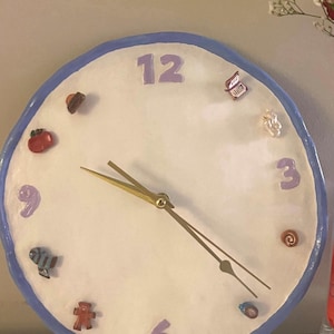 May include: A round wall clock with a white face, a blue border, and purple numbers 12, 3, 6, and 9. The clock features small, colourful, three-dimensional decorative charms representing various objects between the numbers and has two gold-coloured metal hands.
