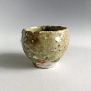 May include: A small, handcrafted ceramic cup with a textured, irregular surface. The cup features a blend of earthy tones, including beige, green, and hints of red. The rim is uneven, adding to its rustic charm. The cup is likely used for drinking.