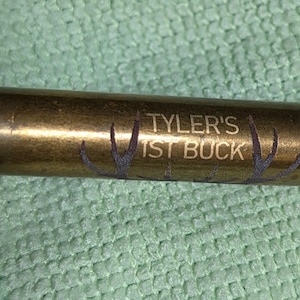 Puede incluir: Una vaina de bala de color latón con el texto "Tyler's 1st Buck" y un diseño de cornamenta grabado en el lateral. La vaina está sobre una superficie texturizada de color verde claro.