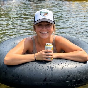 Puede incluir: Una gorra tipo trucker blanca y negra con un parche gráfico de un castor nadando en un río. La gorra presenta un panel frontal blanco estructurado, una visera negra y una parte trasera de malla negra con un diseño de un castor en el agua.