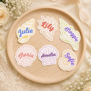 May include: Six die-cut stickers shaped like seashells, each featuring a unique name in cursive script: Julie, Lily, Maggie, Maria, Amelia, and Sofie. The stickers are rendered in pastel colors including yellow, red, green, pink, purple, and orange.