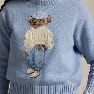 May include: Light blue sweater featuring a detailed embroidered bear wearing a cap, sweater, and jeans. The bear is holding tools. The sweater has ribbed cuffs and hem, with the initials "RL" embroidered on the lower right.
