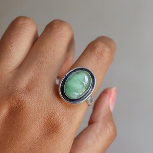 May include: A silver ring featuring a smooth, oval-shaped light green stone set in a dark, oxidized metal bezel. The stone has subtle white marbling throughout its surface, and the ring band is polished silver, creating a contrast with the dark setting.
