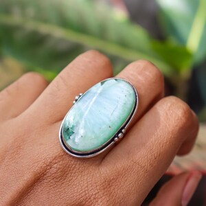 May include: Oval-shaped ring featuring a large, light blue-green gemstone with a silver-colored metal setting. The gemstone has a marbled appearance with darker green inclusions. The ring is set on a finger, showcasing the jewelry's size and design.