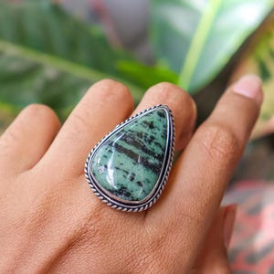May include: A silver ring with a large, teardrop-shaped stone displaying a distinctive green and black pattern. The stone is set within a detailed, twisted silver border. The ring is shown on a hand, with a blurred green and pink background.