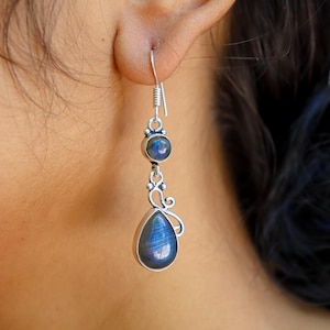 Puede incluir: Pendiente plateado con una piedra preciosa azul en forma de lágrima y una piedra redonda más pequeña. El pendiente tiene un diseño decorativo en espiral y un gancho. Las piedras preciosas son iridiscentes.