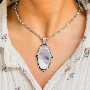 May include: A silver necklace featuring an oval pendant with a milky white stone containing dark, tree-like dendritic inclusions. The pendant is framed by a decorative silver border with small bead-like accents and hangs from a silver chain.