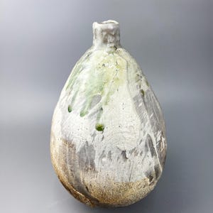 May include: A handmade ceramic vase with a rounded form and a slender neck. The vase has a textured finish with a mix of white, grey, and green tones, and a band of tan near the base. The vase has a rustic, organic appearance.