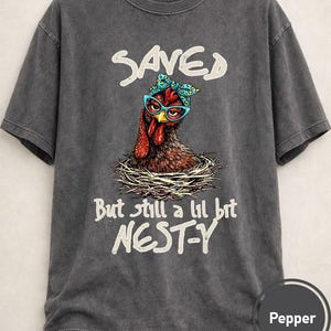 May include: Dark gray t-shirt featuring a whimsical illustration of a chicken wearing glasses and a bow. The text above reads "SAVED", and below, "But still a lil bit NEST-Y". The shirt is a Pepper color.