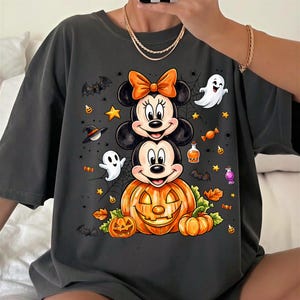 May include: A dark gray t-shirt featuring a graphic print of Minnie and Mickey Mouse stacked on top of a large orange jack-o'-lantern. The design includes small ghosts, bats, stars, potion bottles, and autumn leaves scattered around the characters.