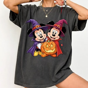 May include: A charcoal gray short-sleeve t-shirt featuring a graphic print of two cartoon mice dressed as witches with purple and red capes and hats. They are standing next to a large orange jack-o'-lantern pumpkin with a small spider nearby.