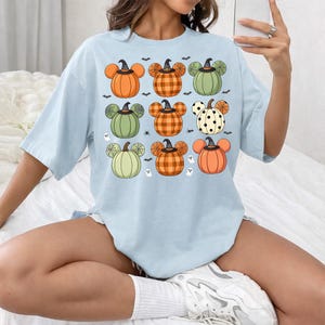 May include: A light blue t-shirt featuring a graphic print of nine pumpkin shapes arranged in a grid. Each pumpkin has mouse-ear silhouettes, some with plaid, polka dot, or solid patterns, and small witch hats. Small bats and ghosts surround them.
