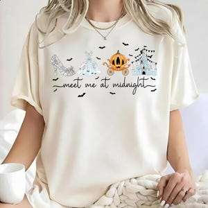 May include: A cream-colored short-sleeve t-shirt featuring a graphic print of a glass slipper, a ballgown, a pumpkin carriage, and a castle. The text "meet me at midnight" is printed in black script below the icons, surrounded by small black bats.