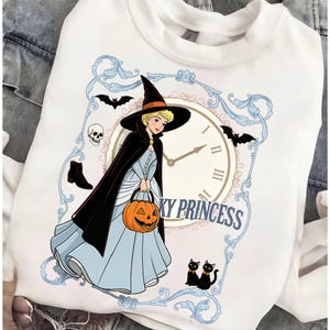 May include: A white sweatshirt featuring a graphic of a blonde character in a black witch hat and cape, holding an orange jack-o'-lantern. The design includes a clock face, bats, a skull, a boot, two black cats, and the text KY PRINCESS.