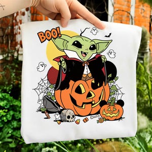 May include: A white t-shirt featuring a graphic of a green creature dressed as a vampire sitting on a large orange jack-o'-lantern. The design includes the text BOO!, small ghosts, a spider web, a skull, candy corn, and a smaller pumpkin.