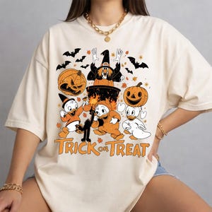 May include: A cream-coloured t-shirt featuring a graphic print of three cartoon ducks in Halloween costumes, a witch in a cauldron, jack-o'-lanterns, bats, and a black cat. The text "TRICK OR TREAT" is printed in orange at the bottom of the design.