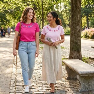 Pode incluir: Duas mulheres caminham por um caminho pavimentado num parque, ambas usando camisetas de manga curta com bordados florais ao redor da gola. Uma &eacute; rosa, a outra rosa claro. Uma mulher usa jeans azuis, a outra uma saia longa bege.
