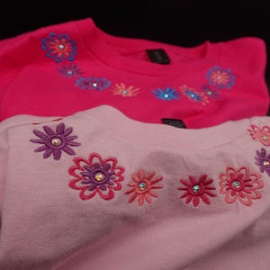 Pode incluir: Duas camisolas rosa com desenhos florais bordados. A camisola superior &eacute; f&uacute;csia vibrante, enquanto a inferior &eacute; um tom de rosa mais claro. Cada uma apresenta uma s&eacute;rie de aplica&ccedil;&otilde;es florais coloridas ao redor da gola.