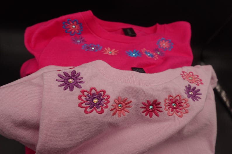 Pode incluir: Duas camisolas rosa com desenhos florais bordados. A camisola superior &eacute; f&uacute;csia vibrante, enquanto a inferior &eacute; um tom de rosa mais claro. Cada uma apresenta uma s&eacute;rie de aplica&ccedil;&otilde;es florais coloridas ao redor da gola.