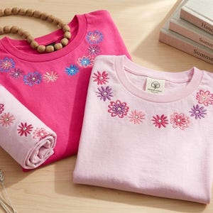Pode incluir: Duas camisetas, uma rosa e outra rosa claro, com desenhos florais bordados ao redor da gola. Uma toalha rosa enrolada com bordados combinando est&aacute; ao lado das camisas. Um colar de contas de madeira e livros tamb&eacute;m est&atilde;o na imagem.