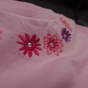 Pode incluir: Uma camisa rosa claro com gola redonda e detalhes florais bordados. As flores s&atilde;o em tons de rosa e roxo, com uma gema prateada no centro de cada flor. A camisa &eacute; feita de um tecido macio.