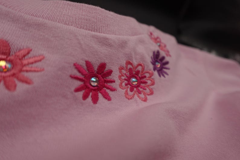 Pode incluir: Uma camisa rosa claro com gola redonda e detalhes florais bordados. As flores s&atilde;o em tons de rosa e roxo, com uma gema prateada no centro de cada flor. A camisa &eacute; feita de um tecido macio.