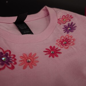 Pode incluir: Camiseta rosa claro com desenhos florais bordados em tons de rosa e roxo. Cada flor tem um centro de strass. A camiseta tem gola redonda e uma etiqueta preta.