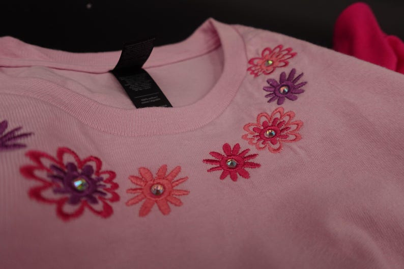 Pode incluir: Camiseta rosa claro com desenhos florais bordados em tons de rosa e roxo. Cada flor tem um centro de strass. A camiseta tem gola redonda e uma etiqueta preta.