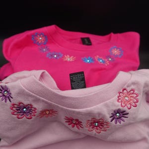 Pode incluir: Duas camisetas rosa com desenhos florais bordados ao redor da gola. Uma &eacute; f&uacute;csia vibrante, a outra em um tom de rosa mais claro. As flores apresentam v&aacute;rias cores e detalhes de strass. Uma etiqueta &eacute; vis&iacute;vel.