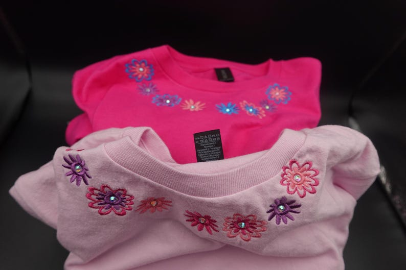 Pode incluir: Duas camisetas rosa com desenhos florais bordados ao redor da gola. Uma &eacute; f&uacute;csia vibrante, a outra em um tom de rosa mais claro. As flores apresentam v&aacute;rias cores e detalhes de strass. Uma etiqueta &eacute; vis&iacute;vel.
