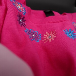 Pode incluir: Uma vibrante camisa rosa com gola redonda adornada com desenhos florais bordados em tons de azul, roxo e p&ecirc;ssego. Cada flor apresenta um adorno brilhante. A camisa &eacute; feita de um material macio.