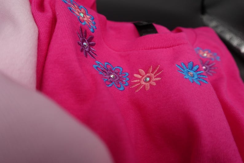 Pode incluir: Uma vibrante camisa rosa com gola redonda adornada com desenhos florais bordados em tons de azul, roxo e p&ecirc;ssego. Cada flor apresenta um adorno brilhante. A camisa &eacute; feita de um material macio.