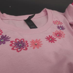 Pode incluir: Uma t-shirt rosa claro com gola redonda e detalhes florais bordados. As flores s&atilde;o em tons de rosa e roxo, cada uma com um strass central. A t-shirt tem uma etiqueta preta na gola.