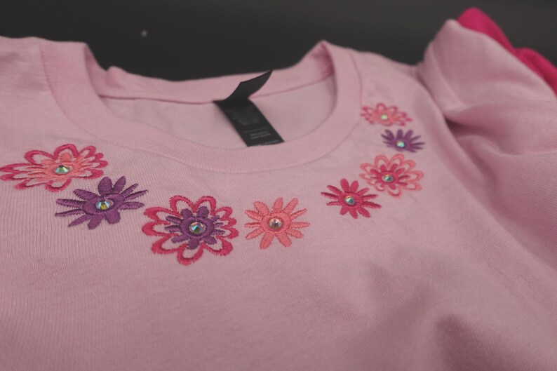 Pode incluir: Uma t-shirt rosa claro com gola redonda e detalhes florais bordados. As flores s&atilde;o em tons de rosa e roxo, cada uma com um strass central. A t-shirt tem uma etiqueta preta na gola.