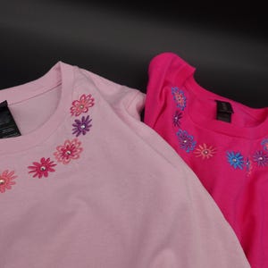 Pode incluir: Duas camisetas de manga comprida, uma rosa claro e outra rosa choque. Ambas as camisas apresentam um design floral ao redor da gola com flores bordadas em v&aacute;rias cores e detalhes de strass. As camisas s&atilde;o feitas de material macio.
