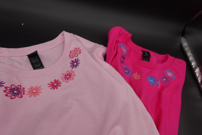 Pode incluir: Duas camisetas de manga comprida, uma rosa claro e outra rosa choque. Ambas as camisas apresentam um design floral ao redor da gola com flores bordadas em v&aacute;rias cores e detalhes de strass. As camisas s&atilde;o feitas de material macio.