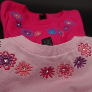 Pode incluir: Duas camisas rosa de manga comprida com desenhos florais bordados. As camisas t&ecirc;m gola redonda e s&atilde;o decoradas com flores em tons de rosa, roxo e azul, cada uma com um strass central. As camisas s&atilde;o provavelmente para crian&ccedil;as.