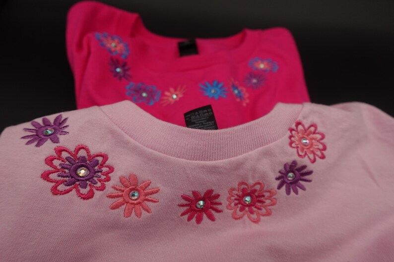 Pode incluir: Duas camisas rosa de manga comprida com desenhos florais bordados. As camisas t&ecirc;m gola redonda e s&atilde;o decoradas com flores em tons de rosa, roxo e azul, cada uma com um strass central. As camisas s&atilde;o provavelmente para crian&ccedil;as.