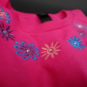 Pode incluir: Uma camisola rosa brilhante com desenhos florais bordados em tons de azul, roxo e coral. Cada flor tem um pequeno enfeite brilhante no centro. A parte superior com gola redonda &eacute; feita de um material macio.