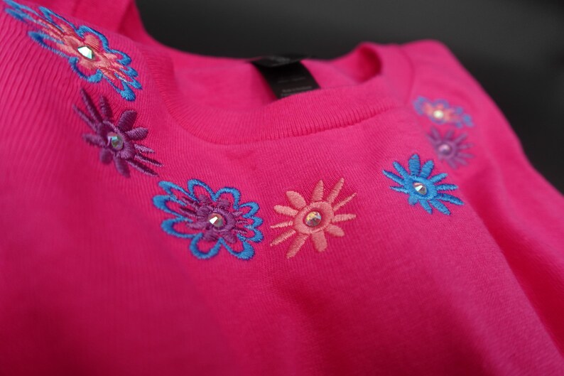 Pode incluir: Uma camisola rosa brilhante com desenhos florais bordados em tons de azul, roxo e coral. Cada flor tem um pequeno enfeite brilhante no centro. A parte superior com gola redonda &eacute; feita de um material macio.