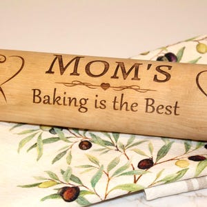 Può includere: Un mattarello di legno con la scritta incisa "MOM'S Baking is the Best". Il mattarello presenta disegni a forma di cuore e floreali a ciascuna estremità. È appoggiato su un canovaccio fantasia con illustrazioni di rami d'ulivo.